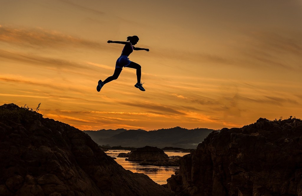 Outline of a runner jumping across a rocky gap at sunset. There is a lake and hills in the background.