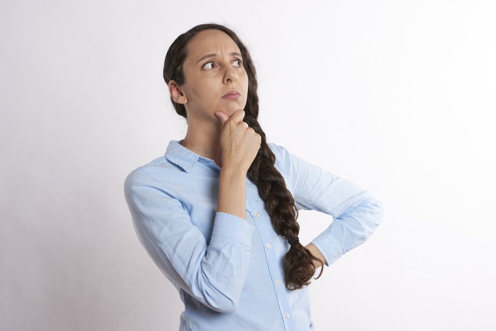 A woman with along brown hair in a plait is wearing a blue shirt and has a thoughtful expression on her face.